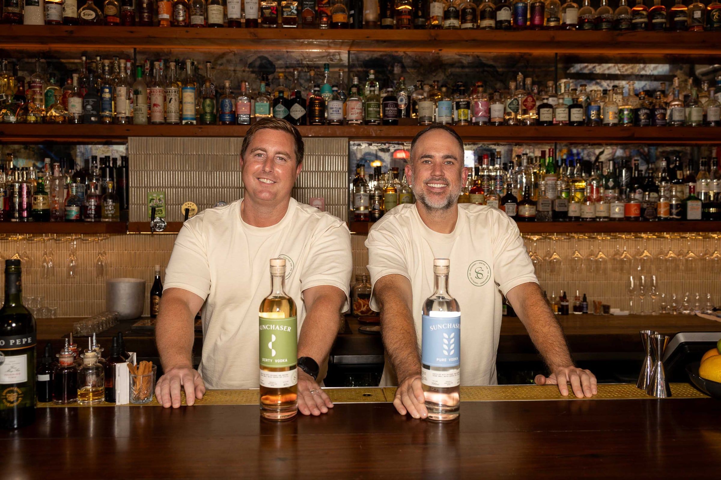 The two Sunchaser founders standing behind a bar with bottles of Dirty and Pure Vodka.