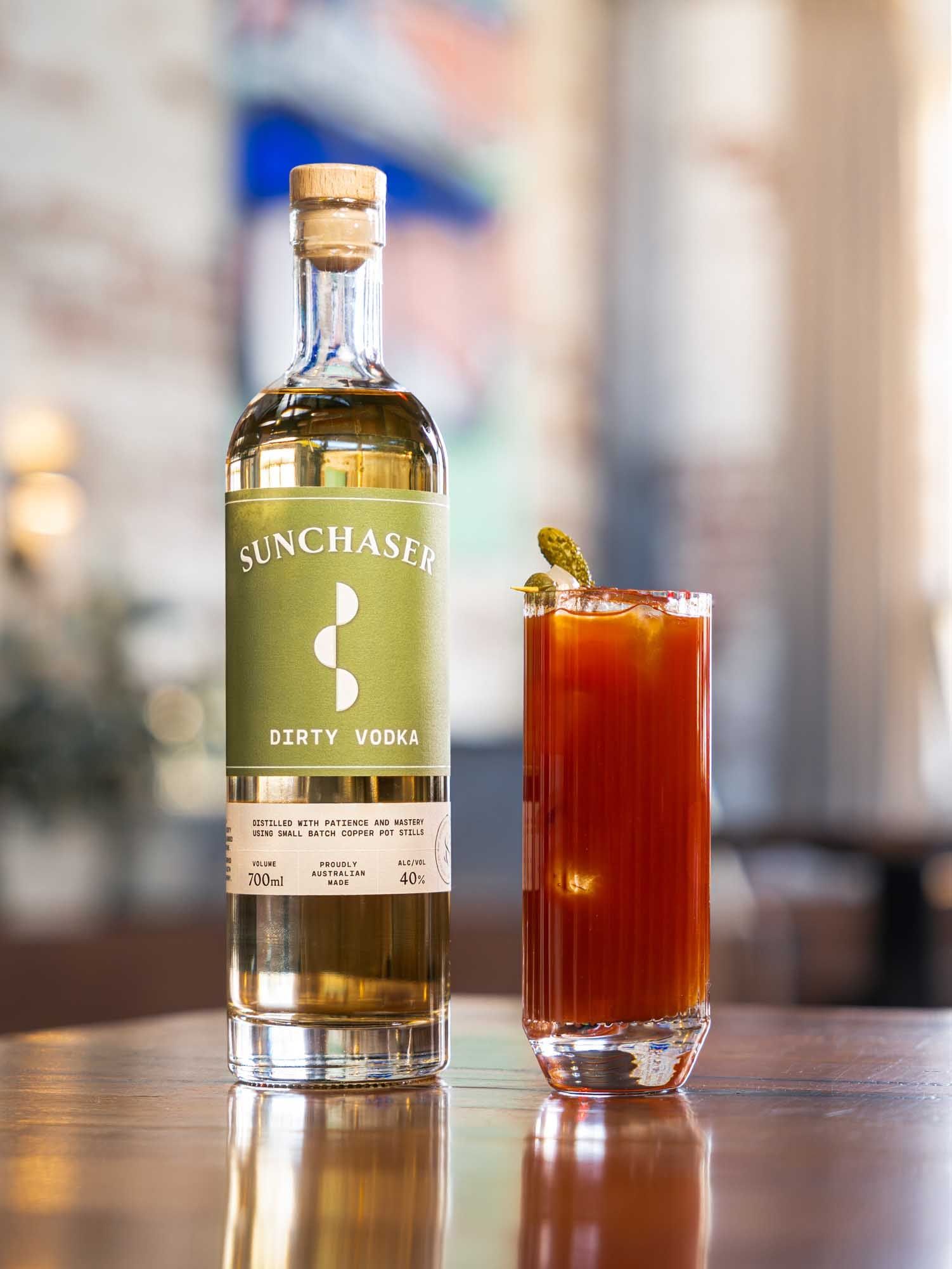 A Bloody Mary in a tall glass beside a bottle of Sunchaser Dirty Vodka on a bar.