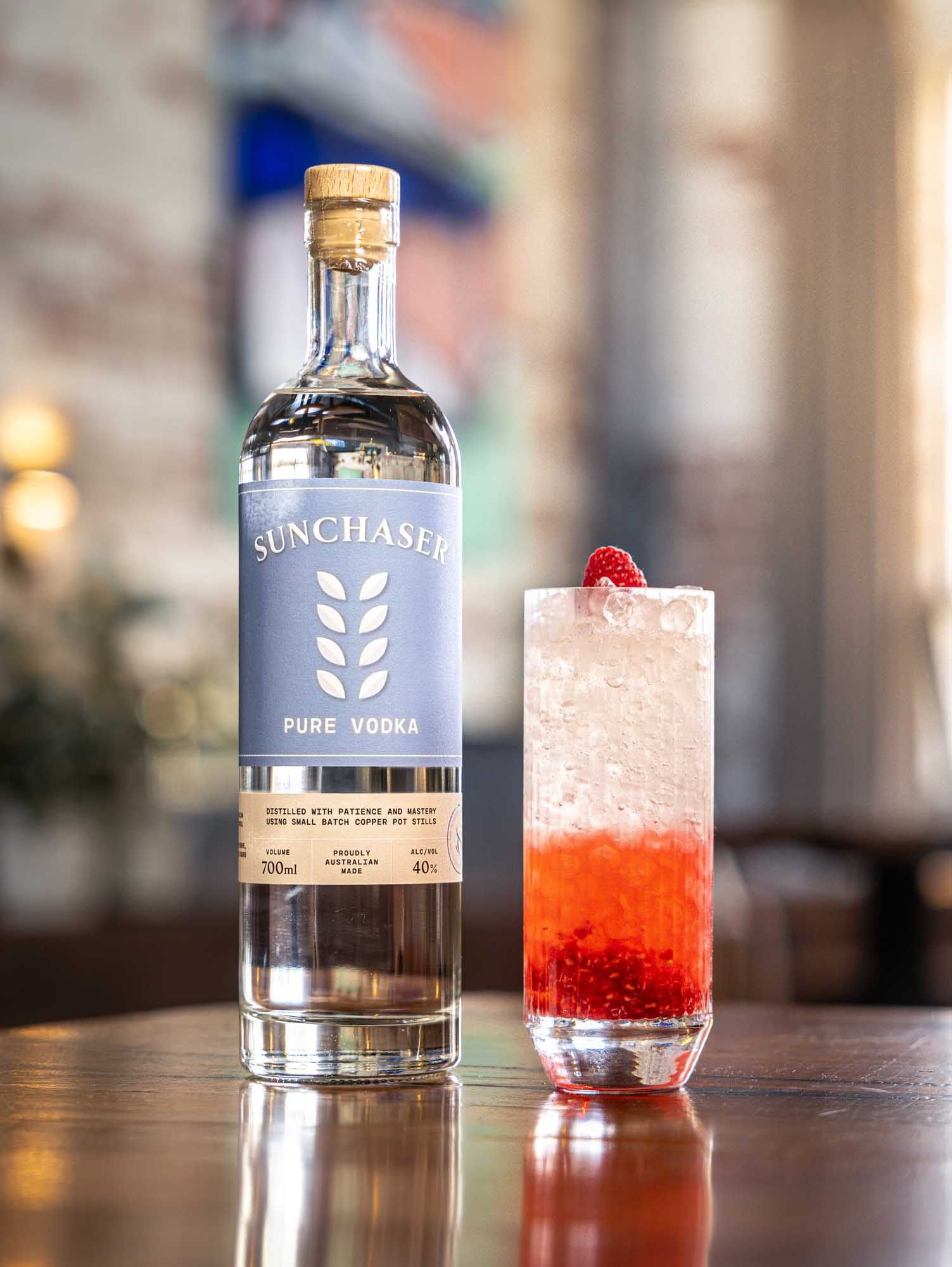 A Spring Punch with a raspberry-red base and a fresh raspberry, beside a bottle of Sunchaser Pure Vodka.